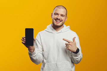 Portrait of happy young man pointing at blank screen mobile phone while smiling, wearing white hoodie, isolated over yellow background.
