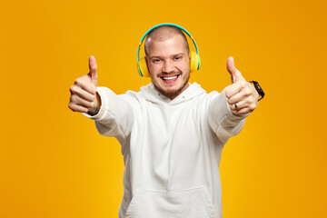 Handsome guy in white hoodie, listening to music in yellow headphones while showing peace gesture with both hands, isolated against orange background