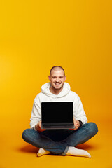 Vertical photo of hipster man wearing white hoodie smiling and showing new laptop while sitting on floor, isolated over yellow background