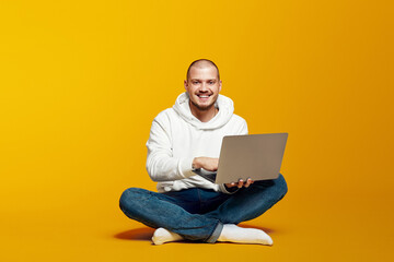Attractive cheerful guy in white hoody sitting lotus pose, using laptop, free time isolated over...