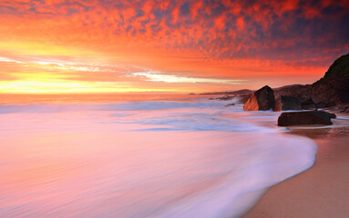Obraz premium Foamy white seas wash onshore and above the skies turn on brilliant hues of yellow to red
