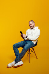 Smiling handsome male using smartphone while sitting on chair isolated on yellow background. Excited casual guy chatting online, browsing social media