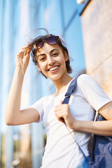 portrait of a young smiling attractive woman in white t-shirt with small city backpack at sunny day on city building background. woman poses in cityscape.