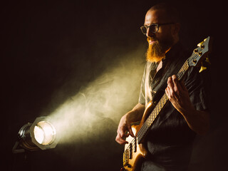 Photo of a bearded man playing bass guitar on stage in front of spotlights.