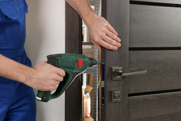 Worker in uniform with screw gun repairing door lock indoors, closeup