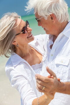 Happy senior man and woman couple dancing and holding hands on a deserted tropical beach with turquoise sea and clear blue sky