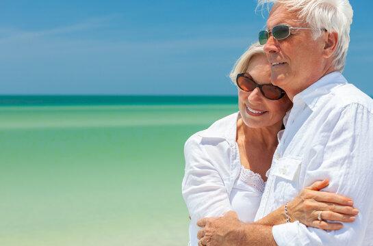 Happy senior man and woman retired couple embracing wearing sunglasses on a deserted tropical beach with turquoise sea and clear blue sky