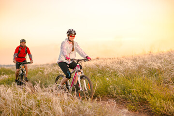 Obraz premium Young Couple Riding the Mountain Bikes in the Beautiful Field Full of Feather Grass at Sunset. Adventure and Family Travel Concept.