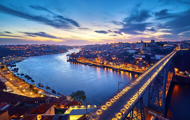 Porto, Portugal. Evening sunset panoramic view at nighttime town and Ponte de Dom Luis bridge with tramways. Coastline of river Douro with reflections of illumination in water and picturesque clouds o