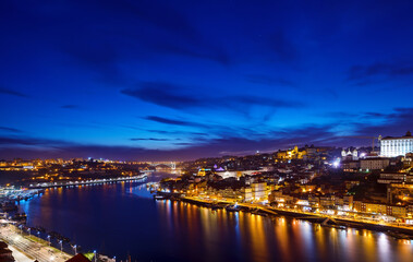 Fototapeta na wymiar Porto, Portugal. Evening sunset panoramic view at nighttime town. Coastline of river Douro with reflections of illumination in water and picturesque clouds on blue sky.