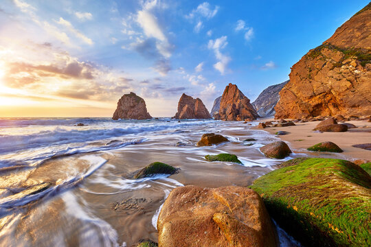 Portugal Ursa Beach at atlantic coast of Atlantic Ocean with rocks and sunset sun waves and foam at sand of coastline picturesque landscape panorama. Stones with green moss in front.