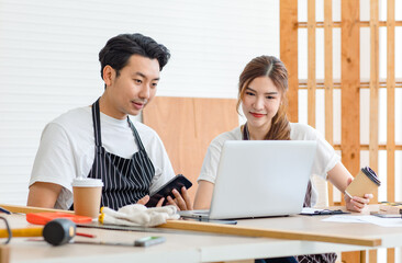 Obraz premium Asian professional male and female lover couple carpenter worker in apron looking pointing blueprint planning via laptop computer on wooden table with equipment in home decoration building workshop