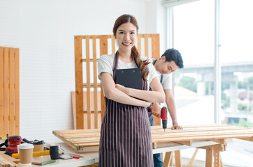 Asian professional female carpenter in apron smiling holding notes on clipboard while male...