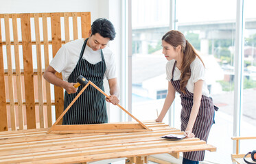 Asian professional male carpenter in apron using rubber hammer pounding on square triangle wood sticks and planks on wooden table workbench while female lover colleague standing looking in workshop