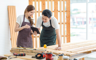 Asian professional male and female carpenter worker lover couple in apron standing smiling helping looking at short notes on paper clipboard together in interior home decoration building workshop
