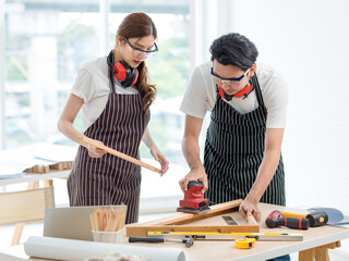 Asian professional skilled male carpenter worker staff in apron with earphones and safety goggles standing holding using polishing machine rubbing polish wooden sticks while female helping in workshop