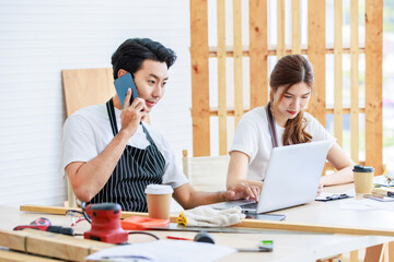 Asian professional male and female lover couple carpenter worker in apron looking pointing blueprint planning via laptop computer on wooden table with equipment in home decoration building workshop