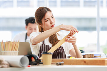 Asian professional focused female carpenter worker staff in apron sitting holding using measuring tape measure wooden sticks on workbench while male lover colleague working on blurred background