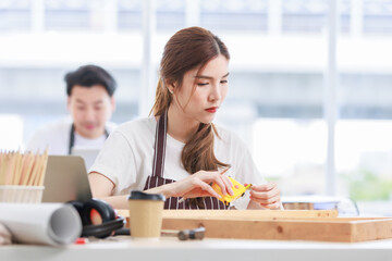 Fototapeta premium Asian professional focused female carpenter worker staff in apron sitting holding using measuring tape measure wooden sticks on workbench while male lover colleague working on blurred background