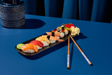A horizontal shot of a sushi set featuring salmon, tuna, shrimp, eel, and avocado. Presented on a black plate against a deep blue background. Modern minimalist Asian cuisine