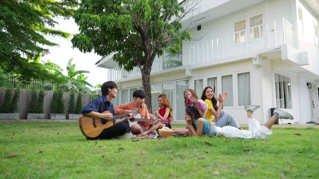Group of Happy young Asian man and woman friends enjoy and fun reunion meeting party at home backyard with playing guitar, singing, eating food and drinking beer together on summer holiday vacation.