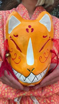 Japanese tradition and beauty: japanese woman in orange silk kimono showing a kitsune mask details under the morning sunlight at serene japanese park