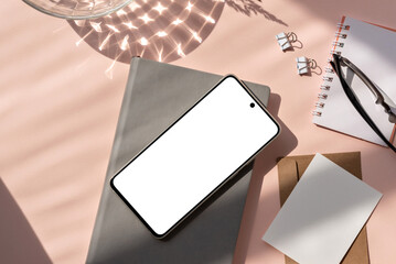 Blank smartphone screen mock up, notebook, paper cards, glasses on pastel pink background with sun light shadow. Minimalist aesthetic stationery flatlay, business brand template, copy space