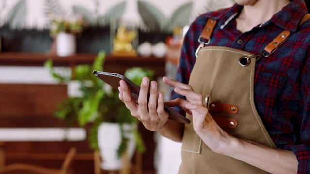 Close-up of beautiful asian woman hands taking orders from customers via delivery app from tablet. It's the first order from the shop. A woman owns a bakery in overalls.