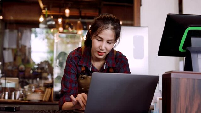 Asian beautiful woman cafe owner Checking orders from customers who ordered cakes and bread. Via online shopping with laptop computer. Teens in apron jumpers look at a store ordering snacks.