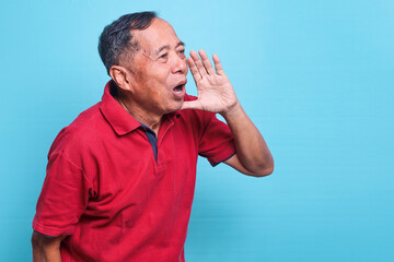 Excited senior Asian man wearing red t-shirt shouting and screaming loud to side with hand on mouth. 