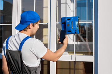 Man in a work jumpsuit and a blue cap takes a picture with a phone of a robot vacuum cleaner of a popular model. A young man remotely turns on the robot through a mobile application