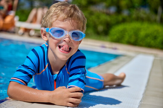 Funny kid in swimming glasses stuck out his tongue lying by the pool with pleasure. Cute Caucasian little boy dabbles playing on the edge of an outdoor pool on a sunny day