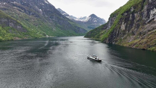 High altitude aerial showing tourism route Hellesylt to Geiranger with ferryboat cruising forward on the Geirangerfjord Norway - Filmed in may month with green forest and snow on mountain peaks