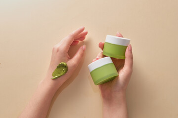 Two green jars is held by a woman hand model and a smear of cream texture on another hand. Mockup of skin care cosmetic jar