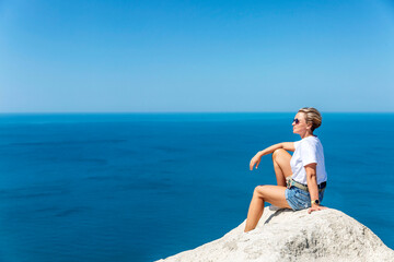 Fototapeta na wymiar A woman in a white tank top and shorts sits on a high white sandy mountain and looks out over the sea and the landscape below against a bright blue sky. Great view. Tourism, travel and recreation.