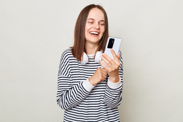 Smiling laughing woman wearing striped shirt standing with headphones over neck isolated over gray background using mobile phone chatting with friends.