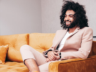 Fototapeta premium Handsome smiling hipster model. Unshaven Arabian man dressed in elegant beige suit . Fashion male with long curly hairstyle posing at home. sitting on yellow sofa
