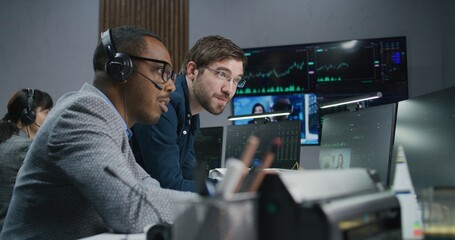 Mixed race stock traders talk and monitor real-time stocks data on computer. Colleagues work in broker agency office. Big digital screen displayed exchange market charts. Investment and analytics.