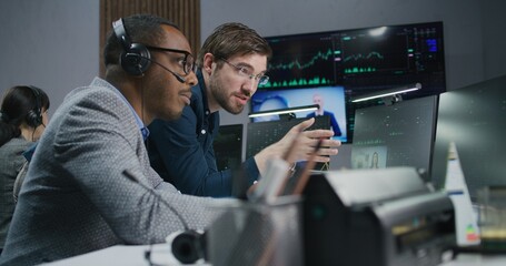 Mixed race stock traders talk and monitor real-time stocks data on computer. Colleagues work in broker agency office. Big digital screen displayed exchange market charts. Investment and analytics.