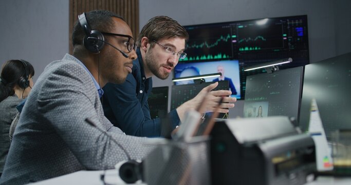 Mixed race stock traders talk and monitor real-time stocks data on computer. Colleagues work in broker agency office. Big digital screen displayed exchange market charts. Investment and analytics.
