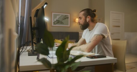 Concentrated man in wireless headphones surfs internet at home. Web designer or developer works...