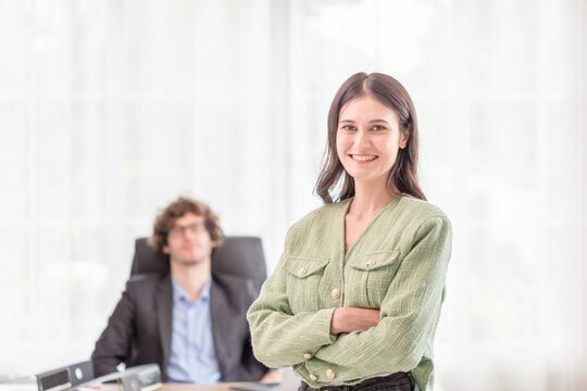 Confident young woman keeping arms crossed and looking at camera with smile while blurred her colleagues in the background