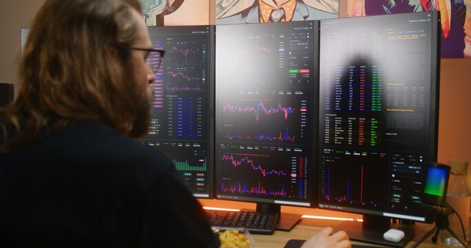Focused male trader analyzes real-time stocks, exchange market charts on multi-monitor computer workstation, eats fast food. Man works remotely in investment at home office. Cryptocurrency trading. - Powered by Adobe