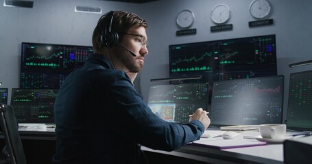 Trader with pen in hand works at computer with displayed real-time stocks. Colleagues analyze exchange market charts on big screens at background. Concept of cryptocurrency trading and investment.