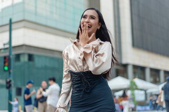 A pretty asian woman standing outside wearing a light tan silk blouse and a high waist skirt, covering her laugh while looking at the sky. A building, people and tents in the background