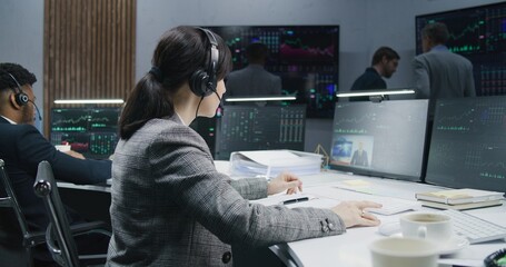 Female trader writes in notepad and works at computer with displayed real-time stocks. Colleagues analyze exchange market charts on big screens at background. Cryptocurrency trading and investment.