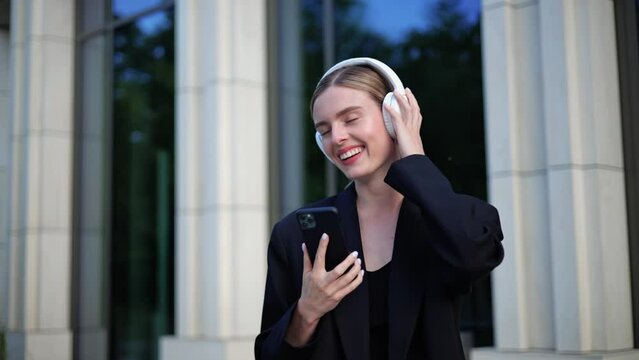 Cool beautiful girl wearing white headphones is listening to music through a new mobile app. Girl is dancing humming along to her favorite song. Woman enjoying a high quality track on the street.