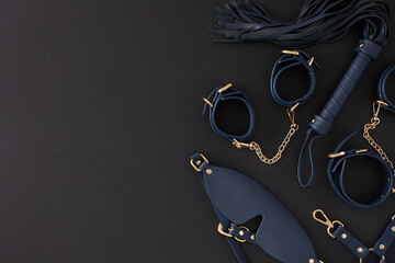 Concept of BDSM toys for sexual satisfaction. Top view composition of handcuffs, black mask, leather whip on black background with empty space for promo or text - obrazy, fototapety, plakaty