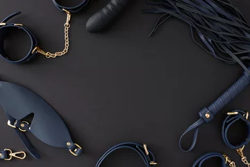 Concept of BDSM toys for erotic indulgence. Top view composition of cuffs, leather mask, whip, dildo on black background with empty space for advert or text © Goncharuk film