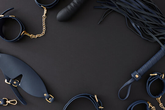 Concept of BDSM toys for erotic indulgence. Top view composition of cuffs, leather mask, whip, dildo on black background with empty space for advert or text